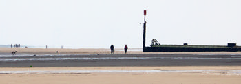 Beach heat haze This landscape photograph captures beach heat haze at Sutton on Sea, located in Lincolnshire, England, United Kingdom. Taken around midday in spring, the image shows people walking along the expansive sandy beach with a visible haze caused by the heat. The beach stretches towards the horizon, where groups of people and a dog can be seen enjoying the shore. Prominently featured is a groyne with a red marker, a typical coastal defence structure found along beaches in the region, providing a recognizable landmark for Sutton on Sea. The soft, diffused light is characteristic of early afternoon in spring, contributing to the humid atmosphere and shimmering visual effect of the heat haze on the Lincolnshire beach.